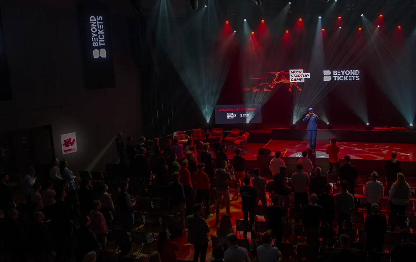 Speaker on the red-lit M01N Startup Camp stage in Rapperswil, the audience standing in front of it, M01N Startup Camp and Beyond Tickets on the screen